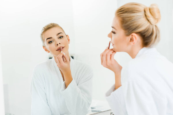 selective focus of beautiful and blonde woman in white bathrobe applying lip liner and looking at mirror 