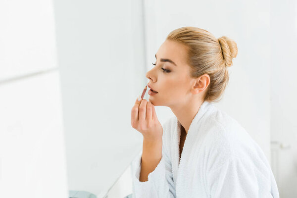 beautiful and blonde woman in white bathrobe applying lip liner in bathroom 