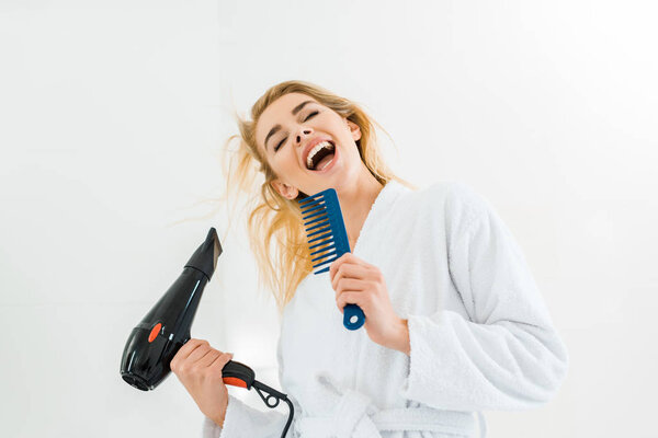 beautiful and smiling woman in white bathrobe singing and holding hairdryer, comb