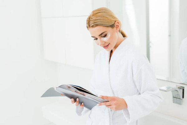beautiful and smiling woman in white bathrobe with eye patches on face reading magazine 