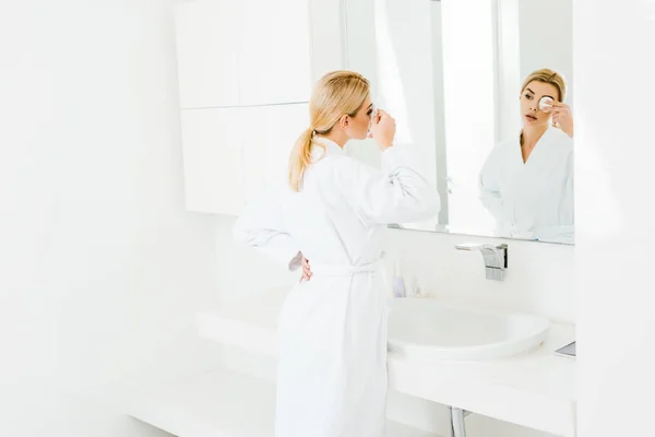 有吸引力的和金发碧眼的女人在白色浴衣使用棉垫和看着镜子的选择性焦点 — 图库照片