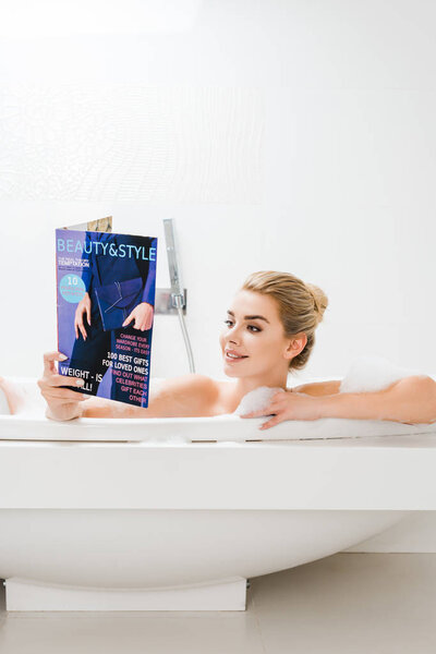 attractive and blonde woman taking bath with foam and reading beauty and style magazine in bathroom 
