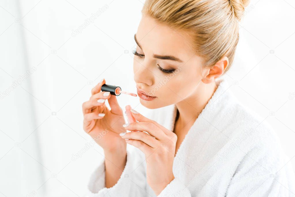 beautiful and blonde woman in white bathrobe applying lip gloss in bathroom 