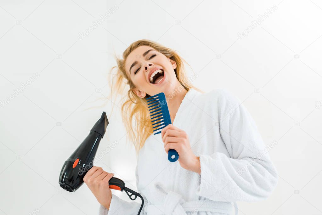 beautiful and smiling woman in white bathrobe singing and holding hairdryer, comb