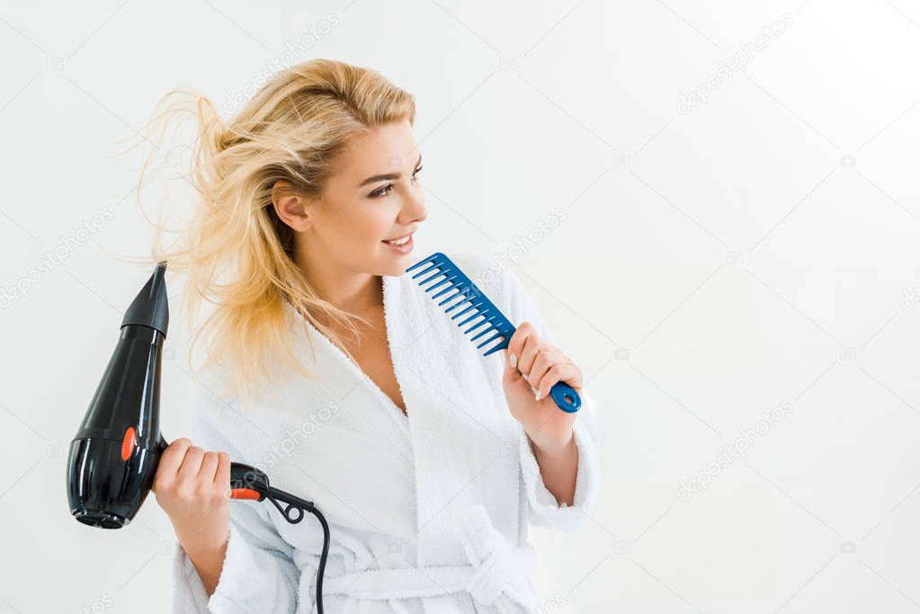 beautiful and smiling woman in white bathrobe holding hairdryer and comb