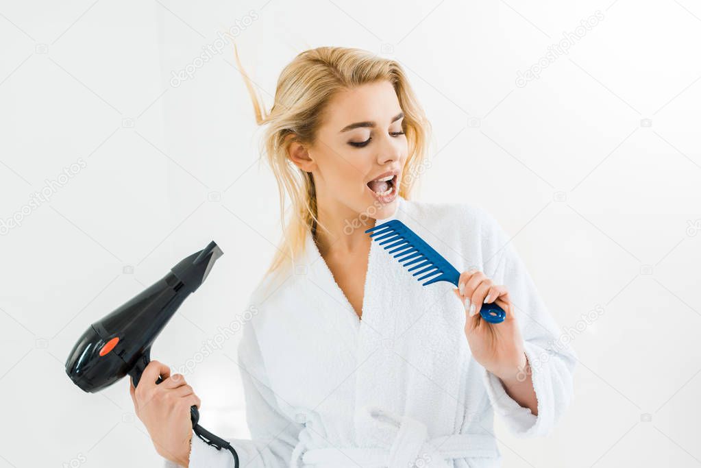 beautiful and smiling woman in white bathrobe singing and holding hairdryer and comb