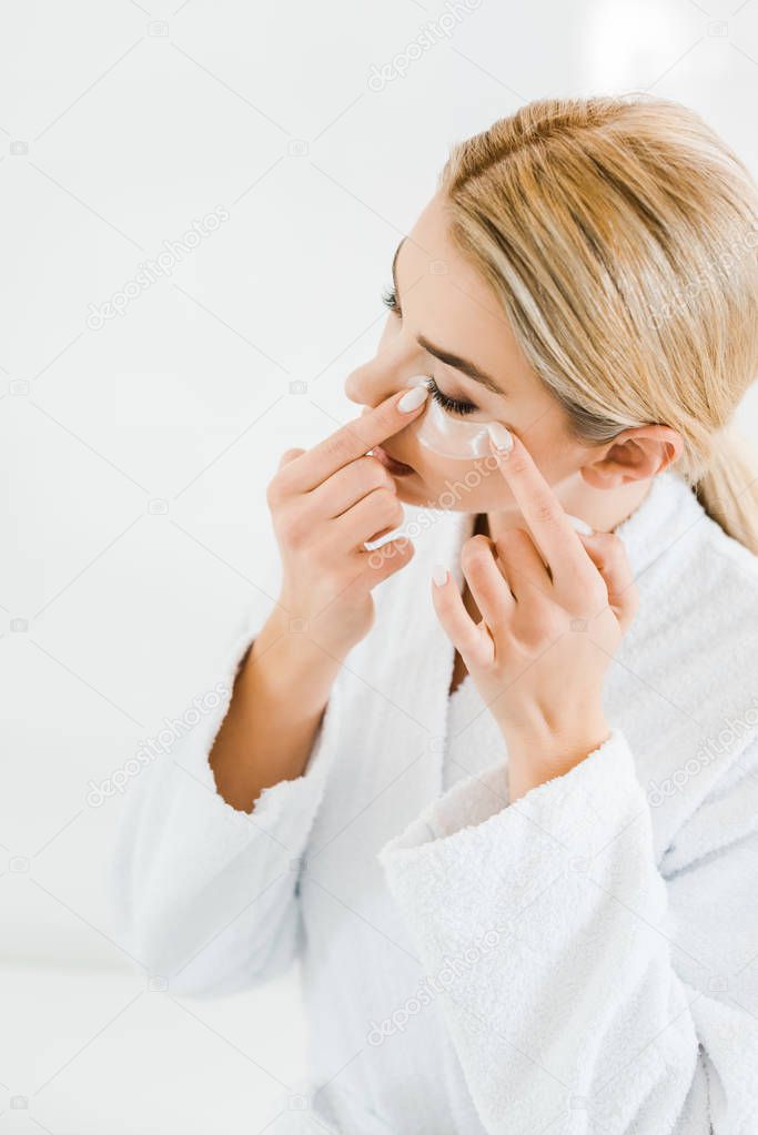 blonde and attractive woman in white bathrobe applying eye patches in bathroom 