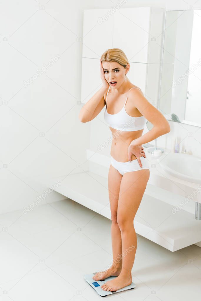 attractive and surprised woman in white bra and panties using scales in bathroom 
