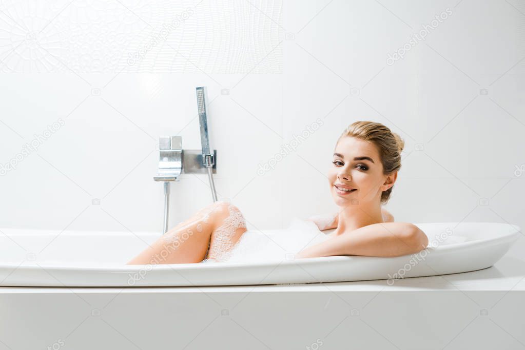 attractive and blonde woman taking bath with foam and looking at camera in bathroom 