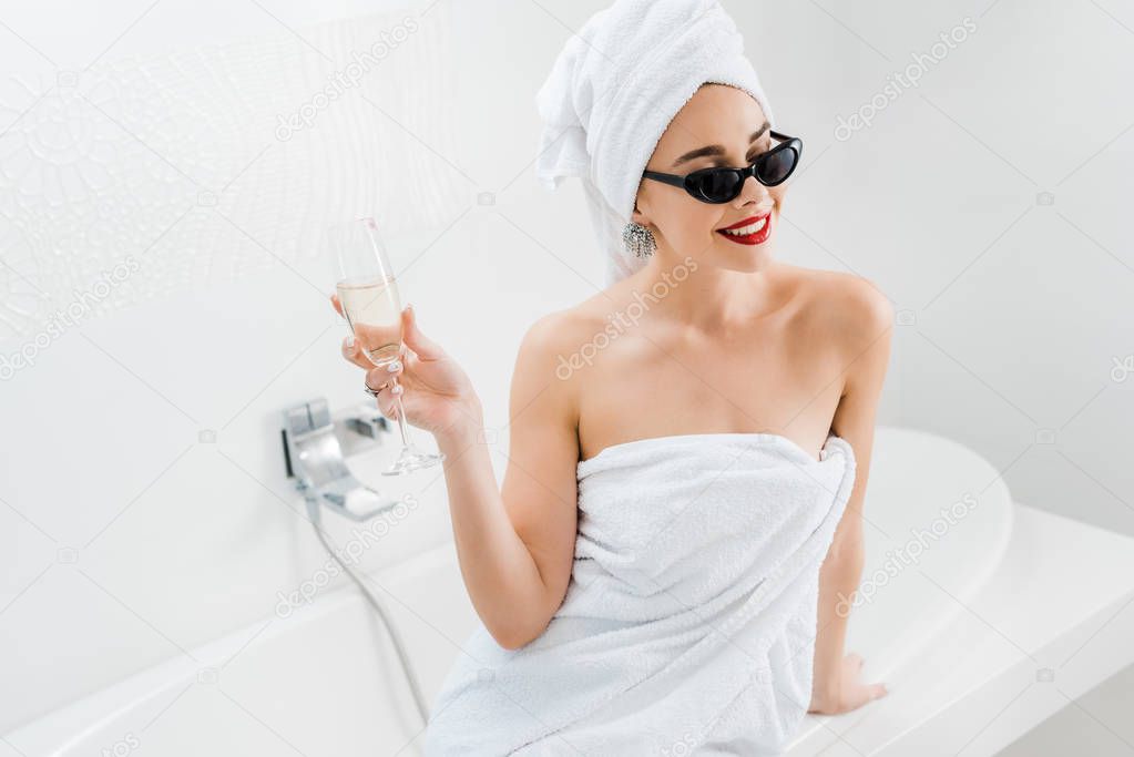 beautiful and smiling woman in sunglasses and towels holding champagne glass in bathroom 
