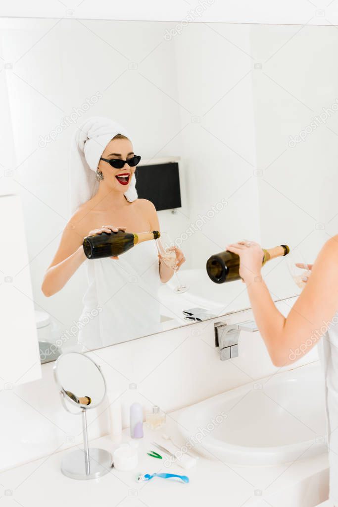 attractive and smiling woman in sunglasses and towels holding champagne glass and bottle in bathroom 