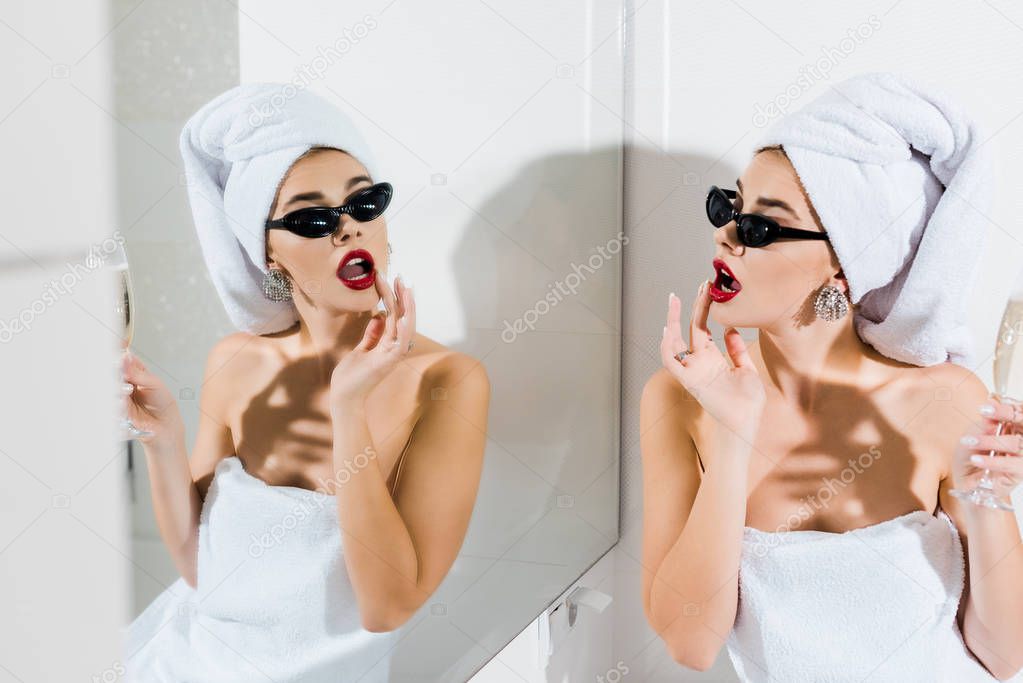 selective focus of attractive woman in sunglasses and towels holding champagne glass and looking at mirror 
