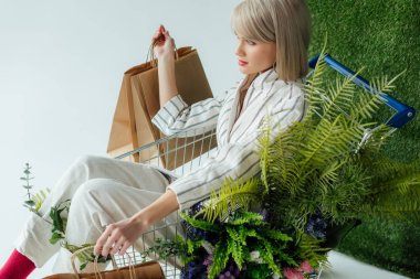 beautiful stylish girl sitting in cart with fern, flowers and shopping bags on white with green grass