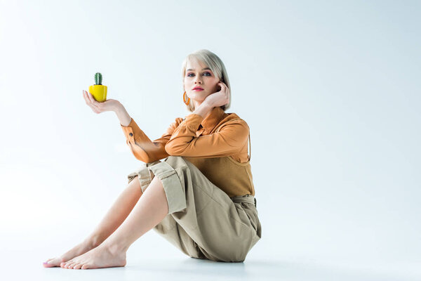 beautiful stylish girl sitting, looking at camera and holding cactus plant isolated on white