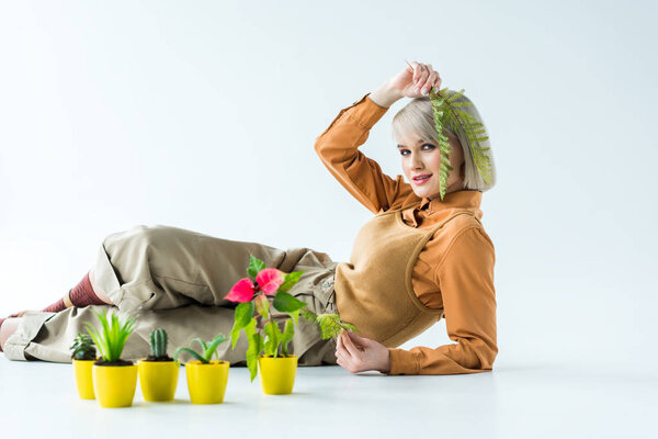 beautiful stylish girl looking at camera and holding fern leaves while posing near flower pots isolated on white