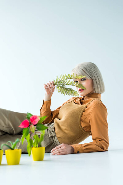 beautiful stylish girl looking at camera and holding fern leaves while posing near flower pots isolated on white