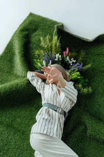 时尚的女孩覆盖脸的手 而躺在人工草与蕨类植物和鲜花 — 图库照片
