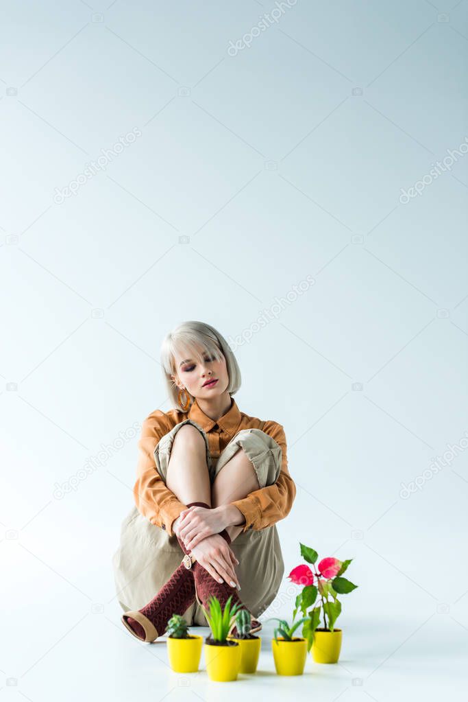 beautiful stylish blonde girl sitting near flower pots on white with copy space