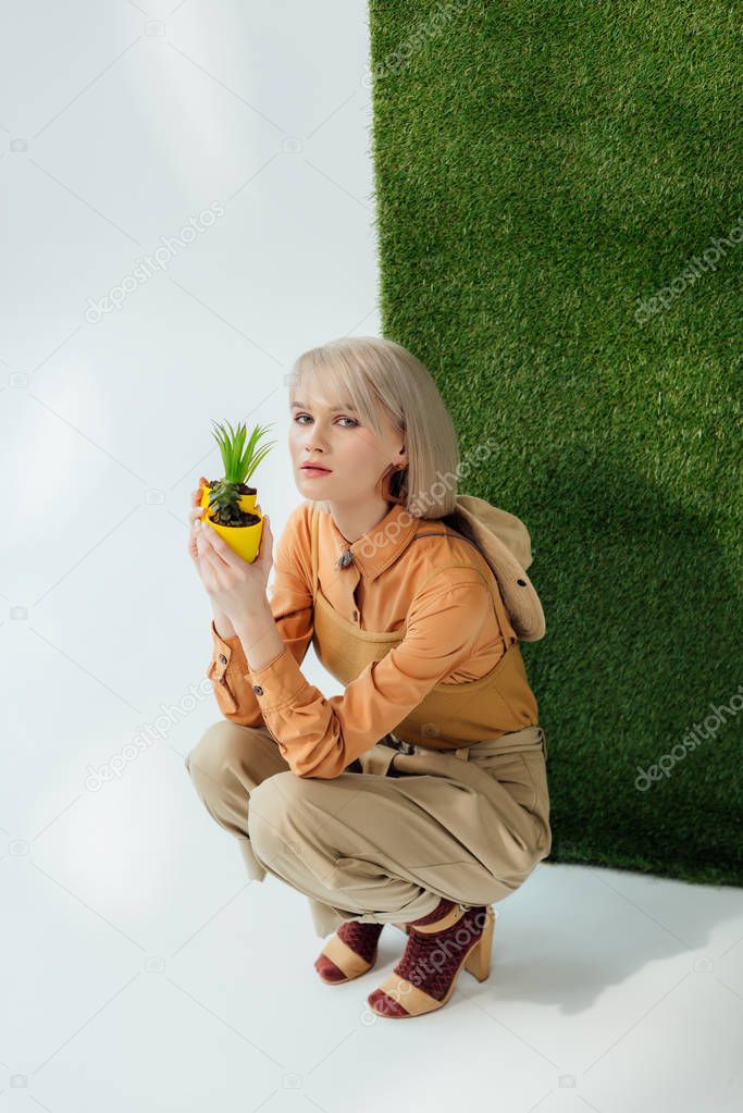 beautiful stylish girl looking at camera while sitting and holding flower pots on grey with green grass 