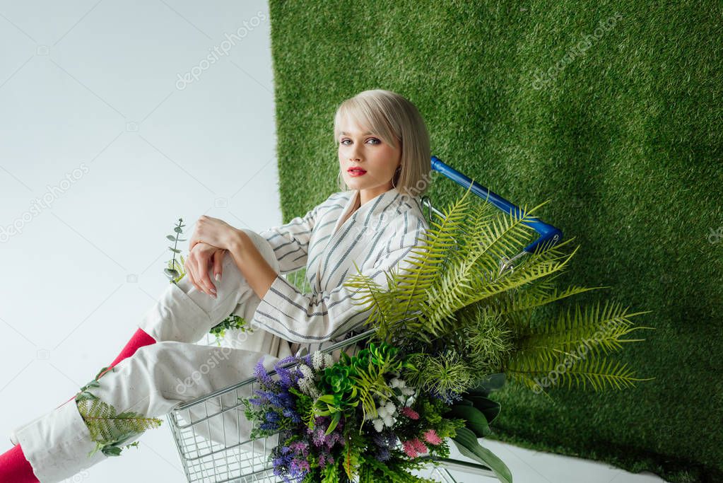 beautiful fashionable girl sitting in shopping cart with fern and flowers on white with green grass