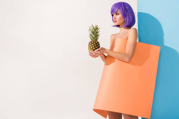 beautiful young woman with purple hair covered in coral paper sheet holding pineapple on blue and white with copy space