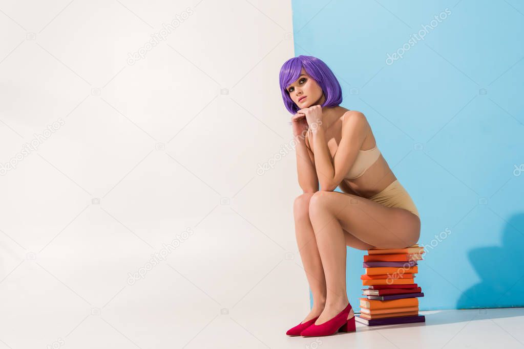 beautiful girl with purple hair looking at camera and sitting on pile of colorful books on blue and white with copy space