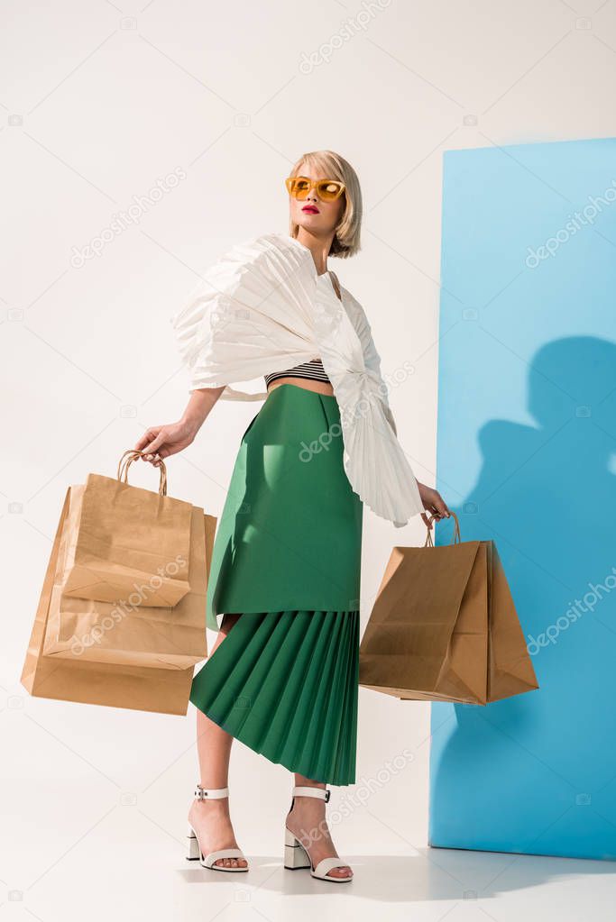 beautiful stylish young woman in sunglasses and paper clothes posing with shopping bags on blue and white