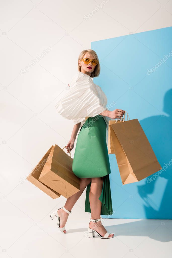 beautiful stylish girl in sunglasses and paper clothes posing with shopping bags on blue and white