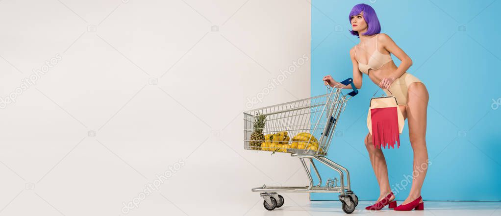 panoramic shot of girl posing near shopping cart with tropical fruits on blue and white with copy space