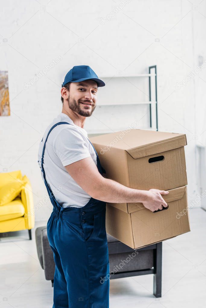 handsome mover in uniform looking at camera while carrying cardboard boxes in apartment