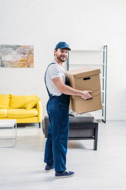 mover in uniform looking at camera while carrying cardboard boxes in apartment clipart