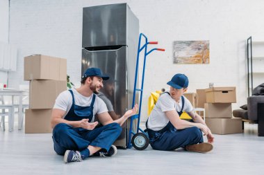 two movers talking while sitting near hand truck, carboard boxes and refrigerator in apartment clipart