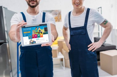cropped view of two movers presenting digital tablet with amazon app on screen in apartment clipart