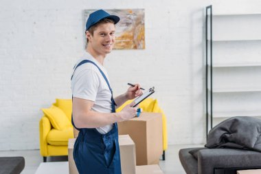 handsome mover holding clipboard and looking at camera near cardboard boxes in apartment clipart