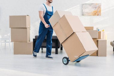cropped view of mover in uniform transporting cardboard boxes on hand truck in apartment clipart