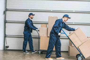 two movers in uniform using hand trucks while transporting cardboard boxes in warehouse clipart