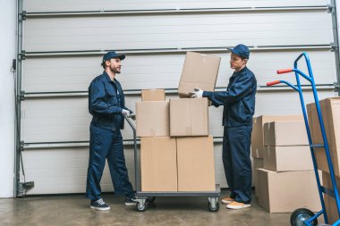 two movers in uniform transporting cardboard boxes on hand truck in warehouse with copy space clipart