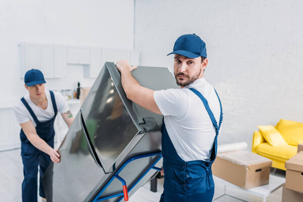 two handsome movers using hand truck while transporting refrigerator in apartment