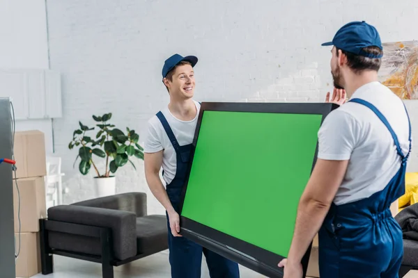 two movers in uniform transporting tv with green screen in apartment