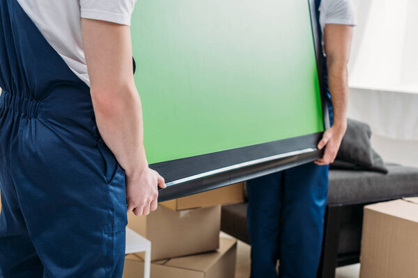 cropped view of two movers in uniform transporting tv with green screen in apartment