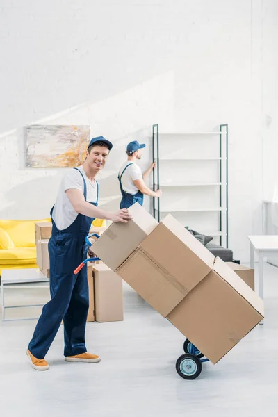 two movers in uniform transporting cardboard boxes and furniture in apartment