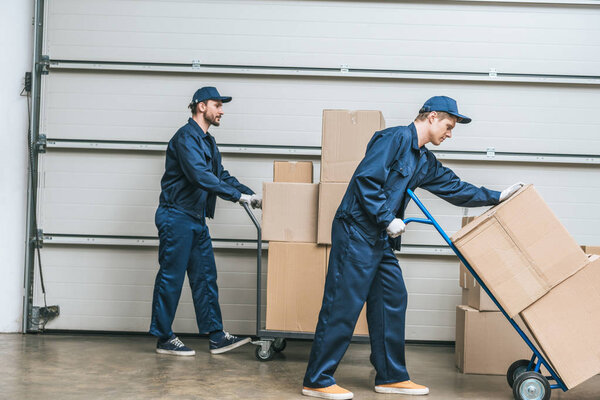 two movers in uniform using hand trucks while transporting cardboard boxes in warehouse
