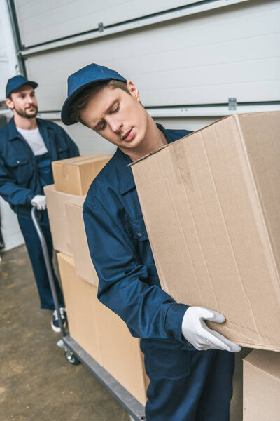 two handsome movers in uniform transporting cardboard boxes with hand truck in warehouse