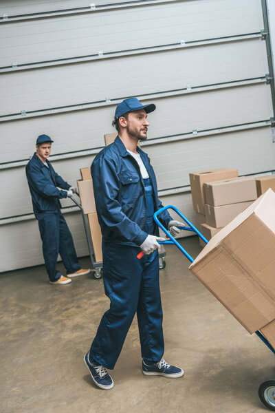 two movers in uniform transporting cardboard boxes with hand trucks in warehouse