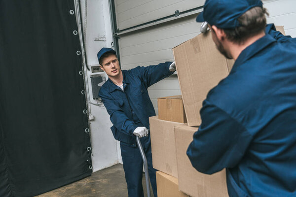 two movers in uniform using hand truck while transporting cardboard boxes in warehouse with copy space