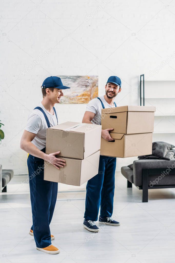 two smiling movers in uniform carrying cardboard boxes in apartment