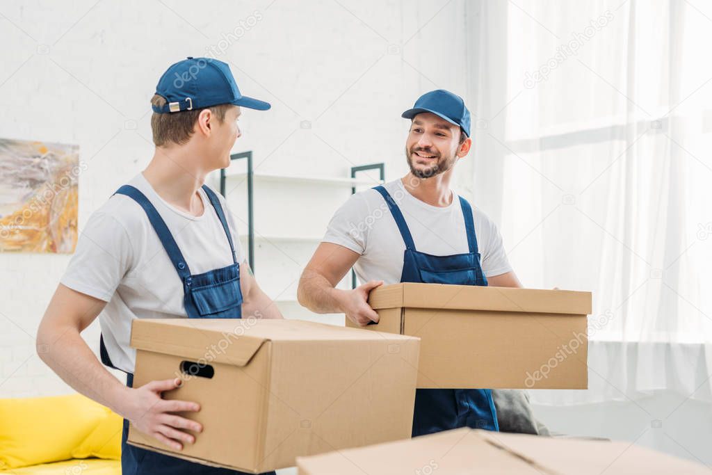 two smiling movers looking at each other while transporting cardboard boxes in apartment