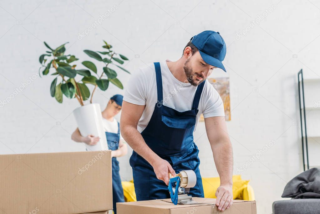 mover in uniform wrapping cardboard box with scotch tape in apartment