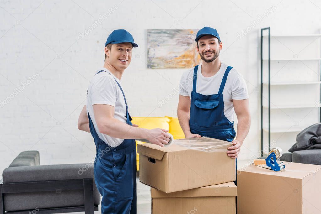 handsome smiling movers looking at camera while wrapping cardboard box with stretch film in apartment