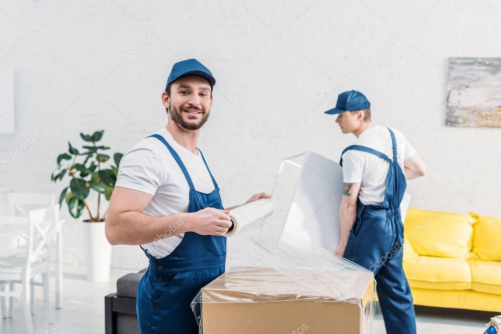 handsome mover in uniform looking at camera while wrapping cardboard box with stretch film in apartment